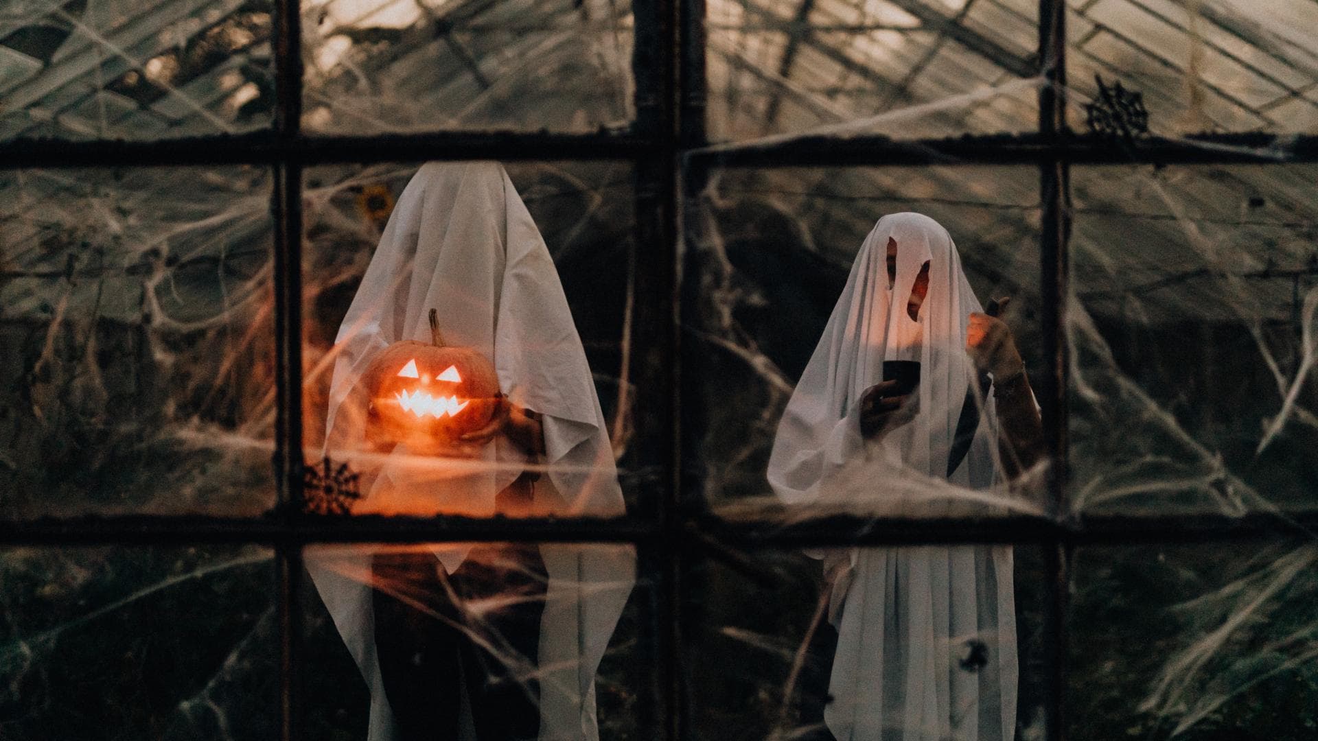 Zwei Menschen als Geist verkleidet, einer hält einen geschnitzten Kürbis in der Hand