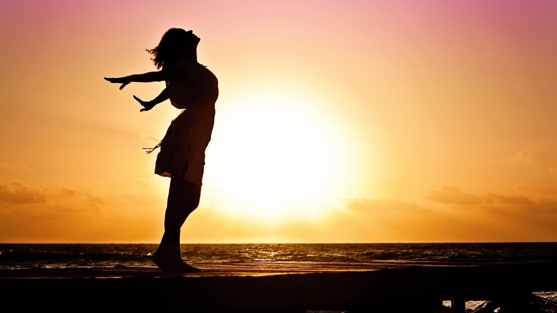 Silhouette einer Frau vor einem Sonnenaufgang, die die Arme nach hinten ausstreckt