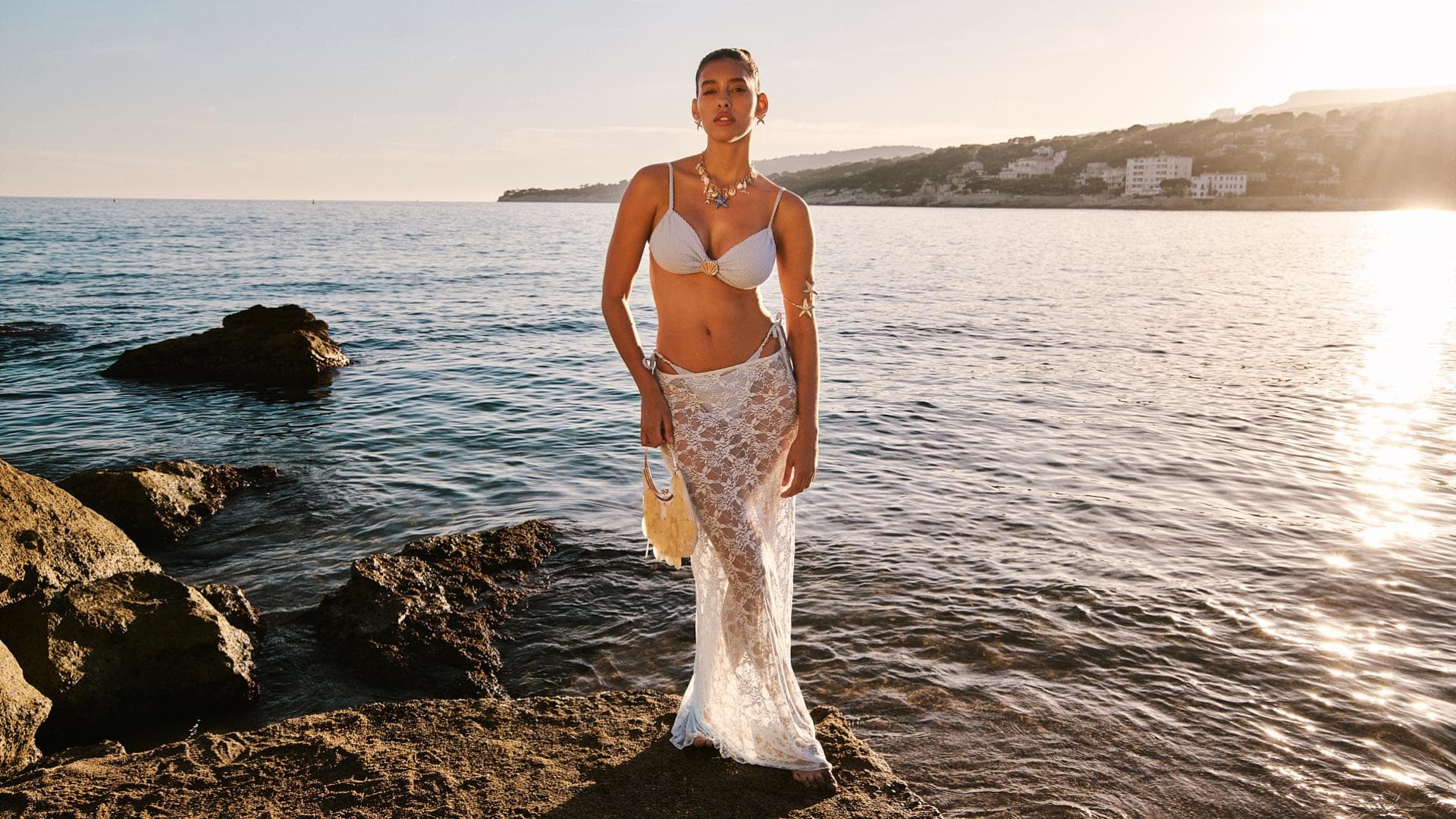Eine junge Frau steht auf einem Felsen im Wasser und trägt einen weißen Bikini und einen Spitzenrock darüber.