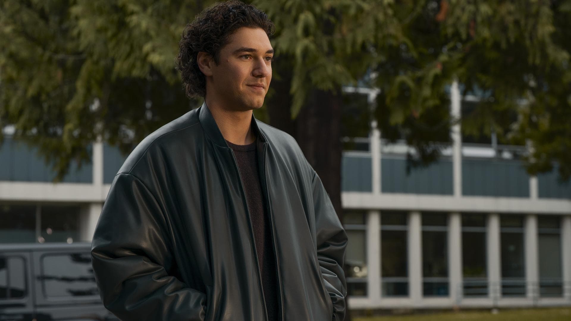Ein junger Mann mit dunklen Locken in einer Lederjacke steht mit seinen Händen in der Tasche und guckt zur Seite.