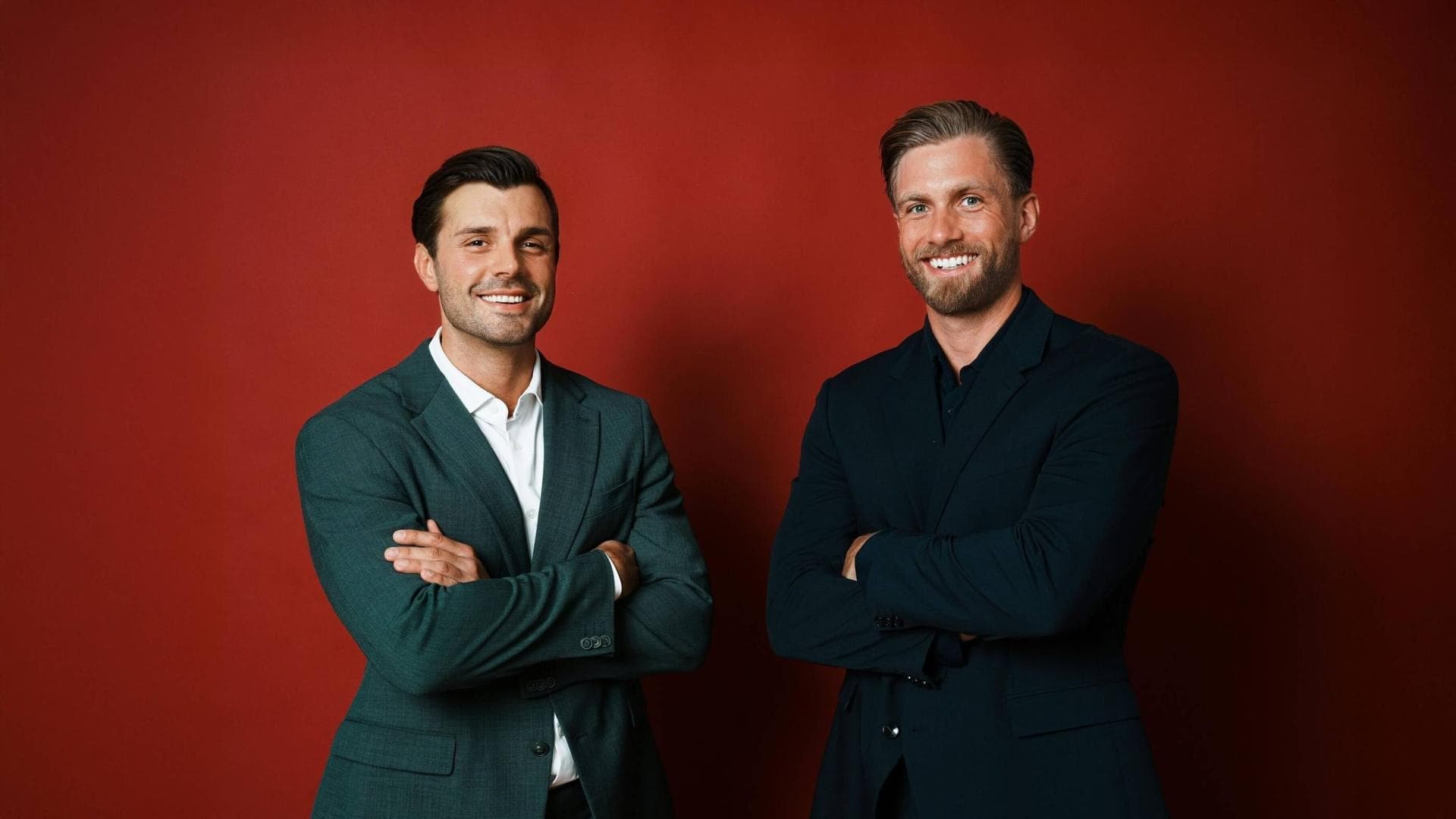 Tim Reitz und Sebastian Paul in Anzügen mit verschränkten Armen vor einer roten Wand.