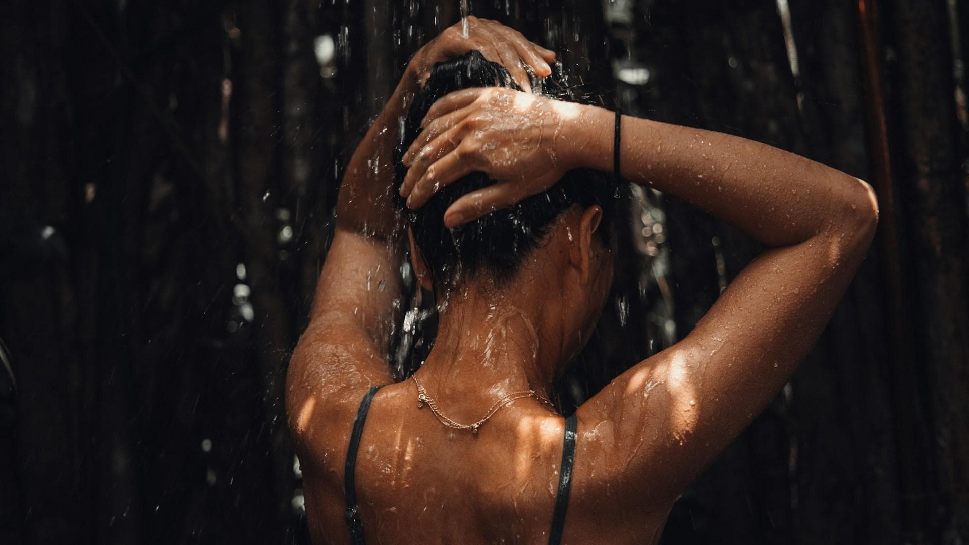 Eine Frau steht mit dem Rücken zur Kamera in einer Dusche und lässt sich mit den Armen über dem Kopf berieseln.