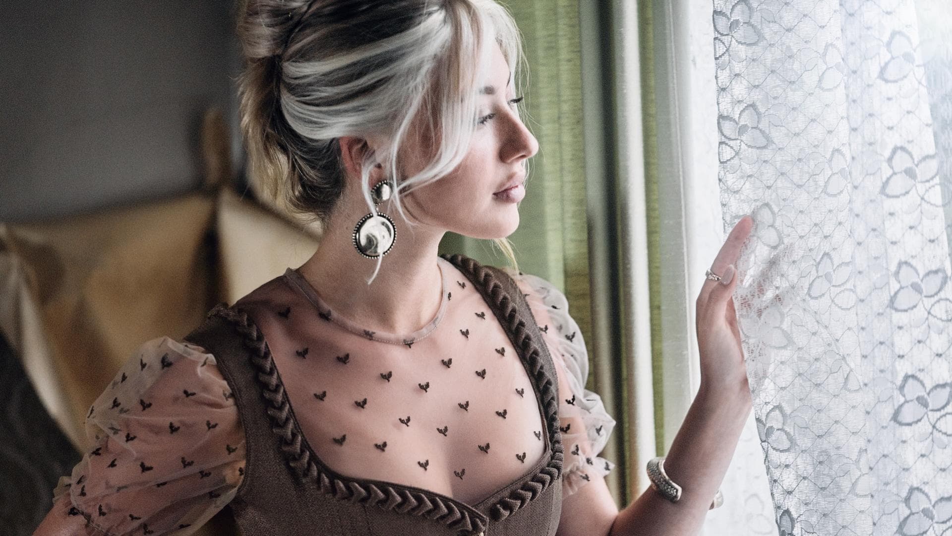 Frau mit hochgesteckten Haaren und Dirndl mit transparenter Bluse guckt durch ein Fester zur Seite.