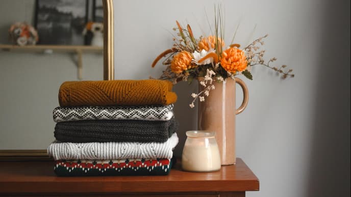 Gefaltete Wollpullover auf einer Holkommode neben einer Kerze und einer Blumenvase