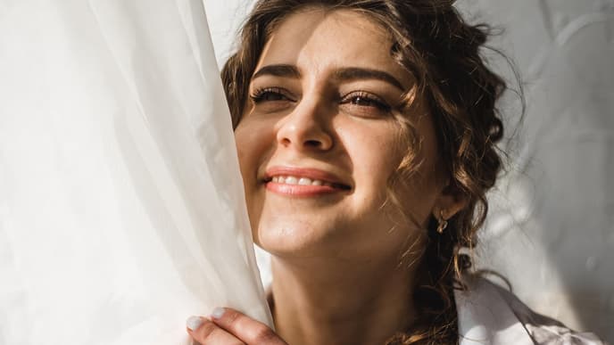 Frau steht hinter weißer Gardine und schaut in die Sonne