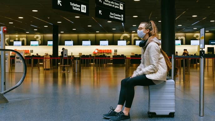 Frau sitzt am Flughafen mit Maske und Koffer