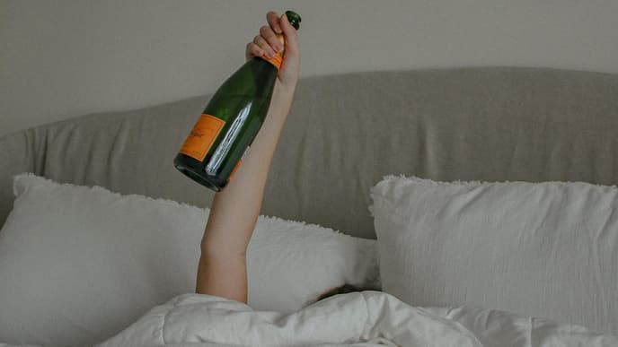 Frau im Bett hält mit einem Arm eine Flasche Alkohol in die Luft