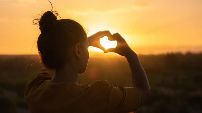 Frau macht Herz mit Händen vor Sonnenuntergang