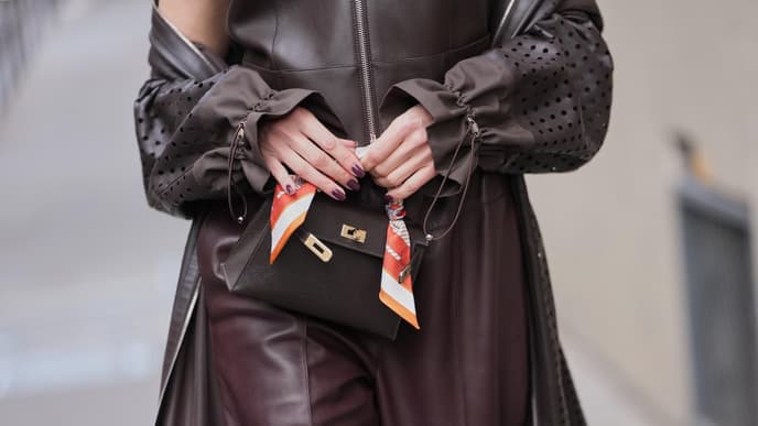 Eine Frau trägt ein braunes Outfit und hält vorn ihre Tasche mit den Fingern fest.