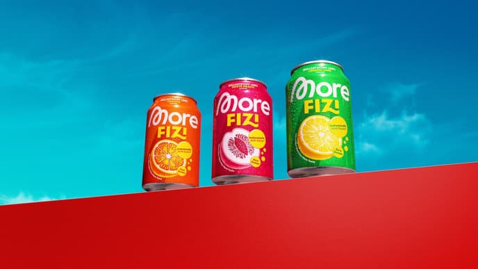 Drei bunte Getränkedosen mit der Aufschrift „More Fizi“ auf einer roten Mauer vor blauem Himmel.