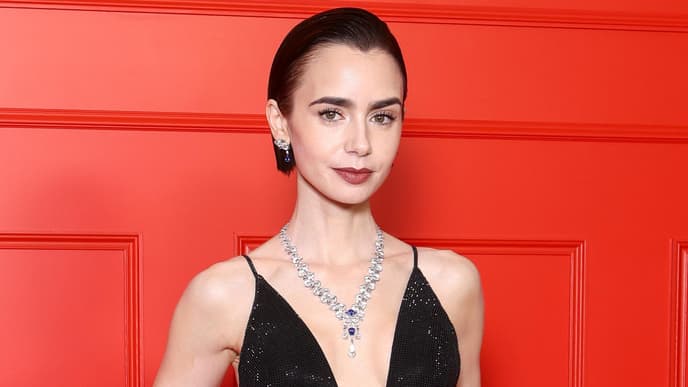 Lily Collins in einem schwarzen Abendkleid mit tiefem V-Ausschnitt vor einer roten Wand.