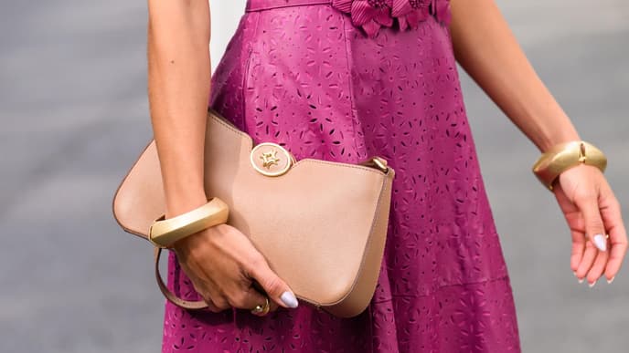 Eine Nahaufnahme einer Frau mit einem Kleid mit Lochmuster in Magenta und einer hellbraunen Clutch in der Hand mit goldenen Armreifen.