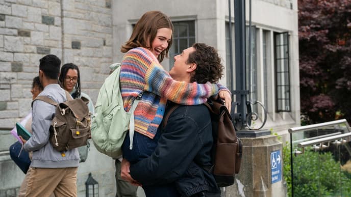 Eine junge Frau mit einem bunt gestreiften Cardigan wird von einem Mann hochgehoben.