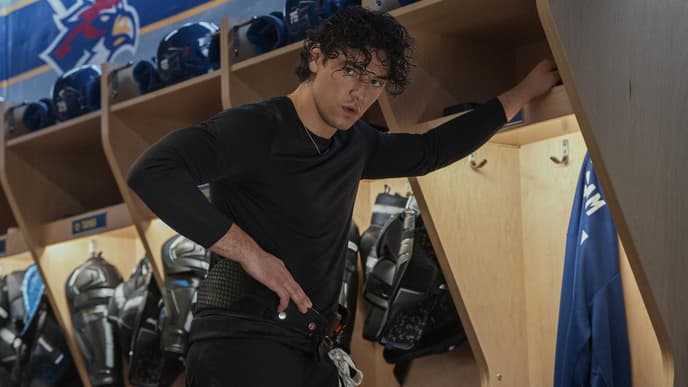 Ein junger Mann mit Locken steht in einem langärmeligen Shirt in der Umkleide einer Eishockey-Mannschaft.