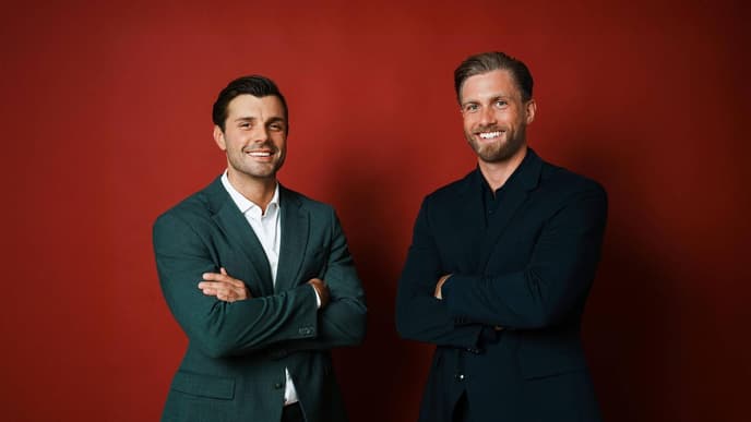 Tim Reitz und Sebastian Paul in Anzügen mit verschränkten Armen vor einer roten Wand.