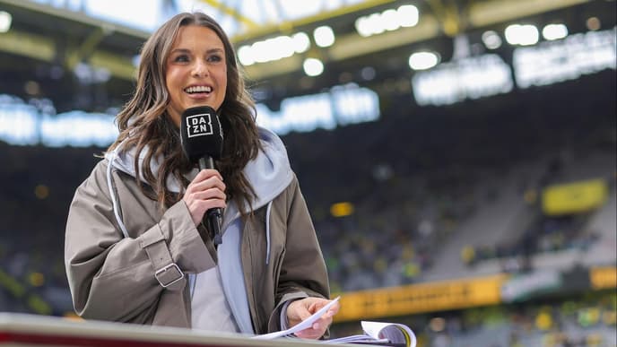 Eine junge Frau mit braunen Haaren steht in einem Fußballstadio und moderiert.