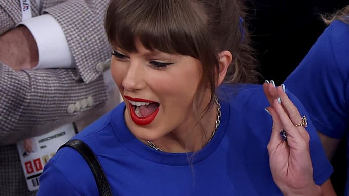 Taylor Swift sorgte mit ihrem Graphic Shirt beim Knicks Spiel für Aufsehen. Taylor Swift mit einem blauen Shirt schreit euphorisch und schaut zur Seite nach links.