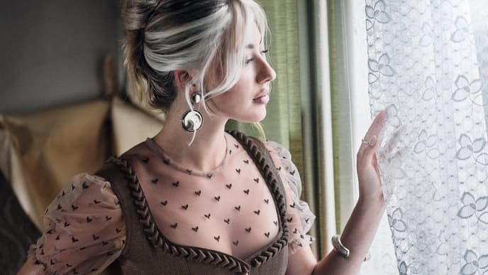 Frau mit hochgesteckten Haaren und Dirndl mit transparenter Bluse guckt durch ein Fester zur Seite.