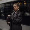 Frau trägt auf Paris Fashion Week braunen Ledermantel goldene Ohrringe und braune Tasche