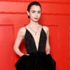 Lily Collins in einem schwarzen Abendkleid mit tiefem V-Ausschnitt vor einer roten Wand.