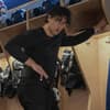 Ein junger Mann mit Locken steht in einem langärmeligen Shirt in der Umkleide einer Eishockey-Mannschaft.