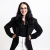 Eine Frau mit schwarzen Haaren, einem schwarzen Blazer und weißer Hose sowie einer Brille mit auf die Hüfte gestemmten Armen.