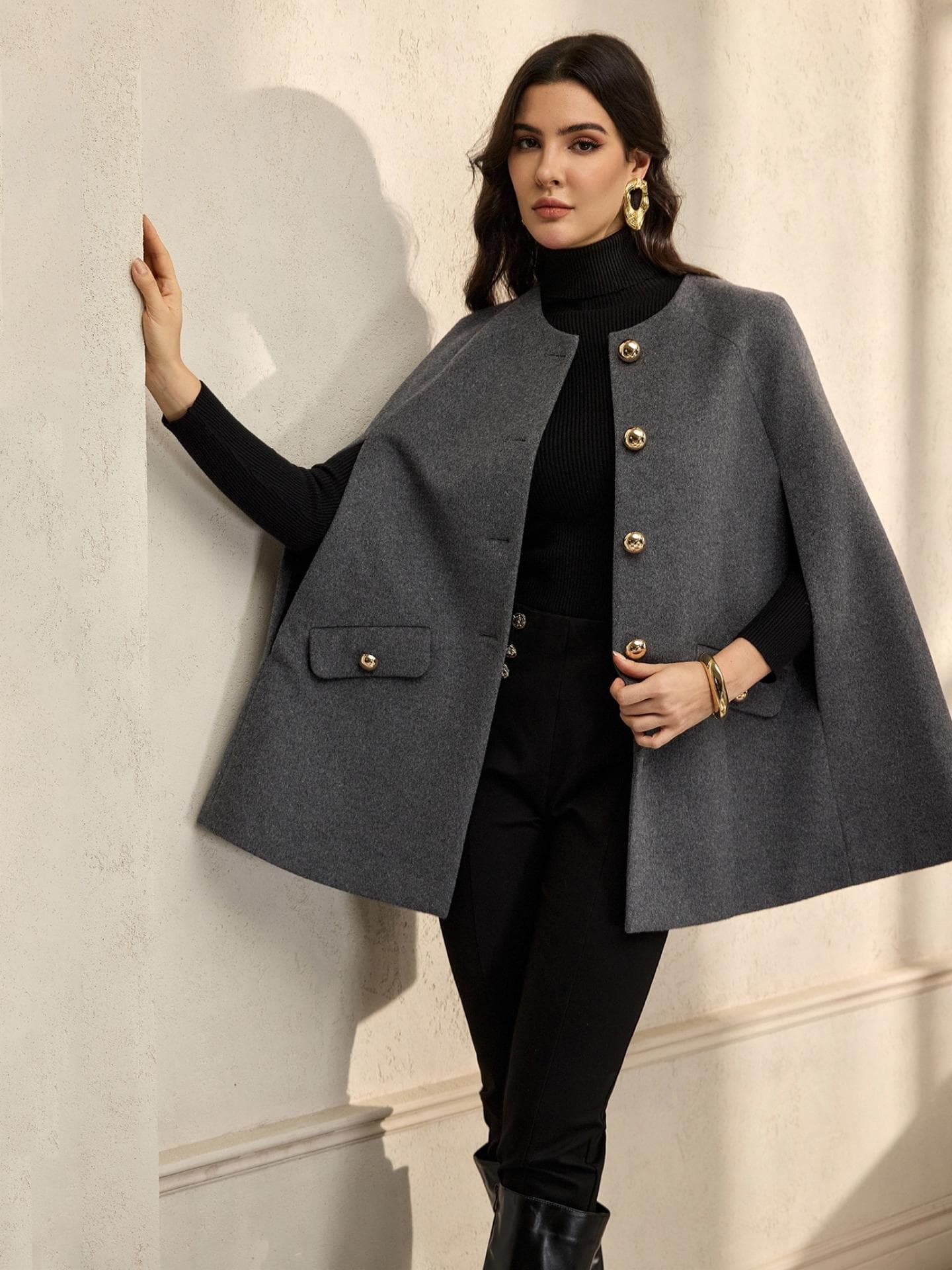 Eine junge Frau mit einem schwarzen Langarmshirt, schwarzer Hose und einem grauen Cape darüber.
