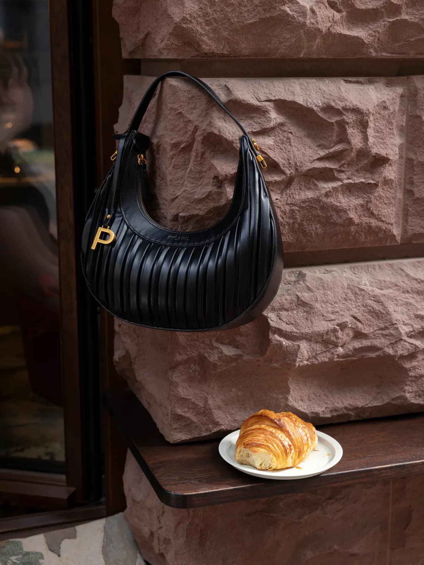 Eine schwarze Schultertasche hängt an einer Wand, darunter steht ein Croissant.