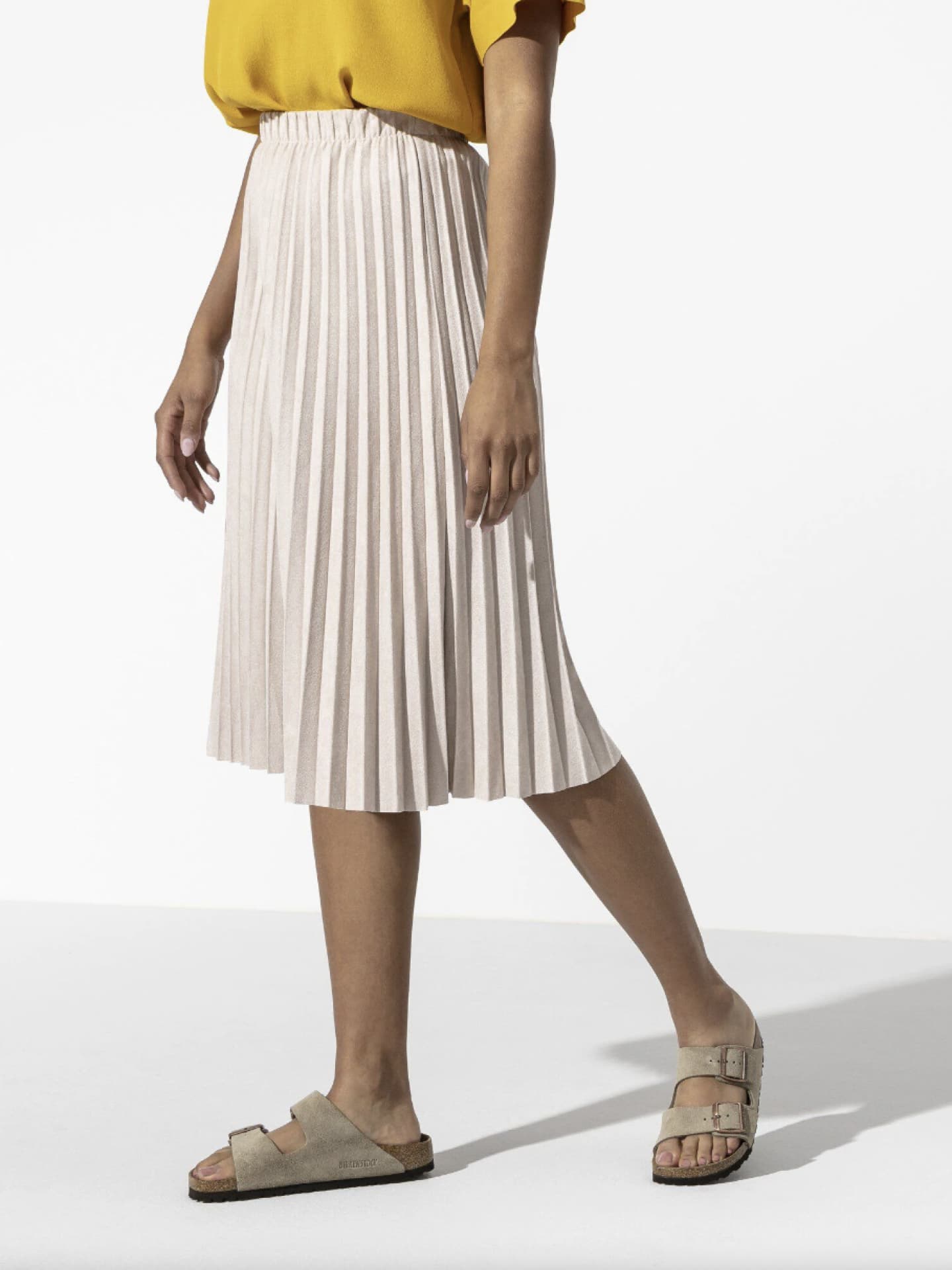 Eine Frau trägt einen Midi-Plisseerock, ein gelbes Oberteil und taupefarbene Birkenstock Sandalen.