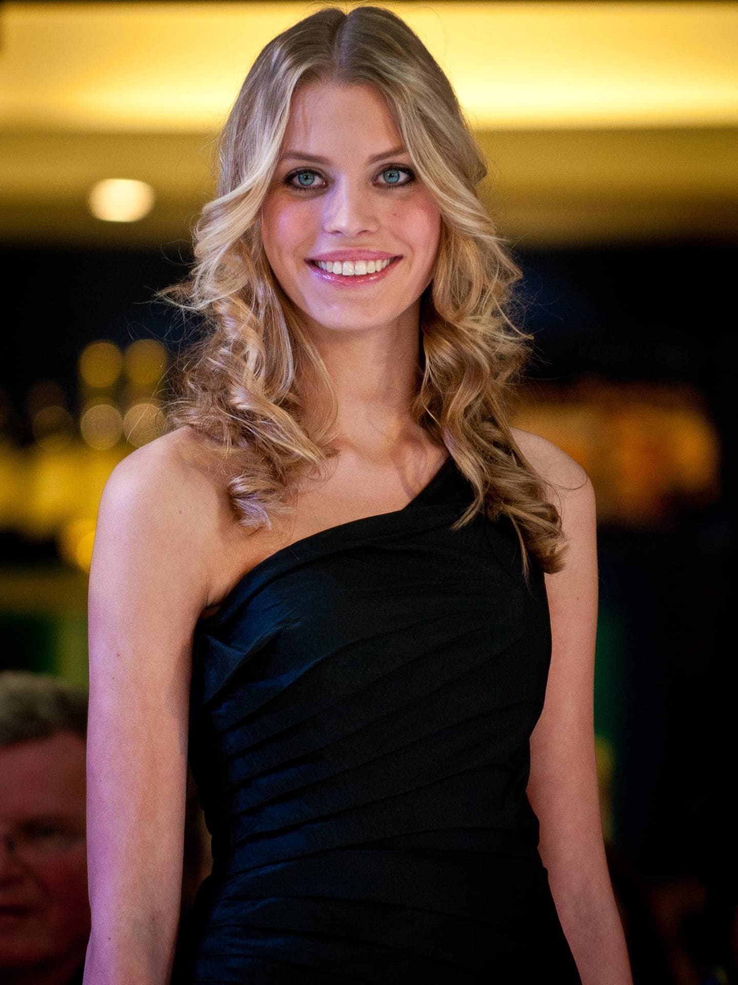 Sarah-Anessa war vor ihrem GNTM-Umstyling blond. Eine junge Frau mit blonden, lockigen Haaren, großen Augen und einem schwarzen One-Shoulder-Kleid.