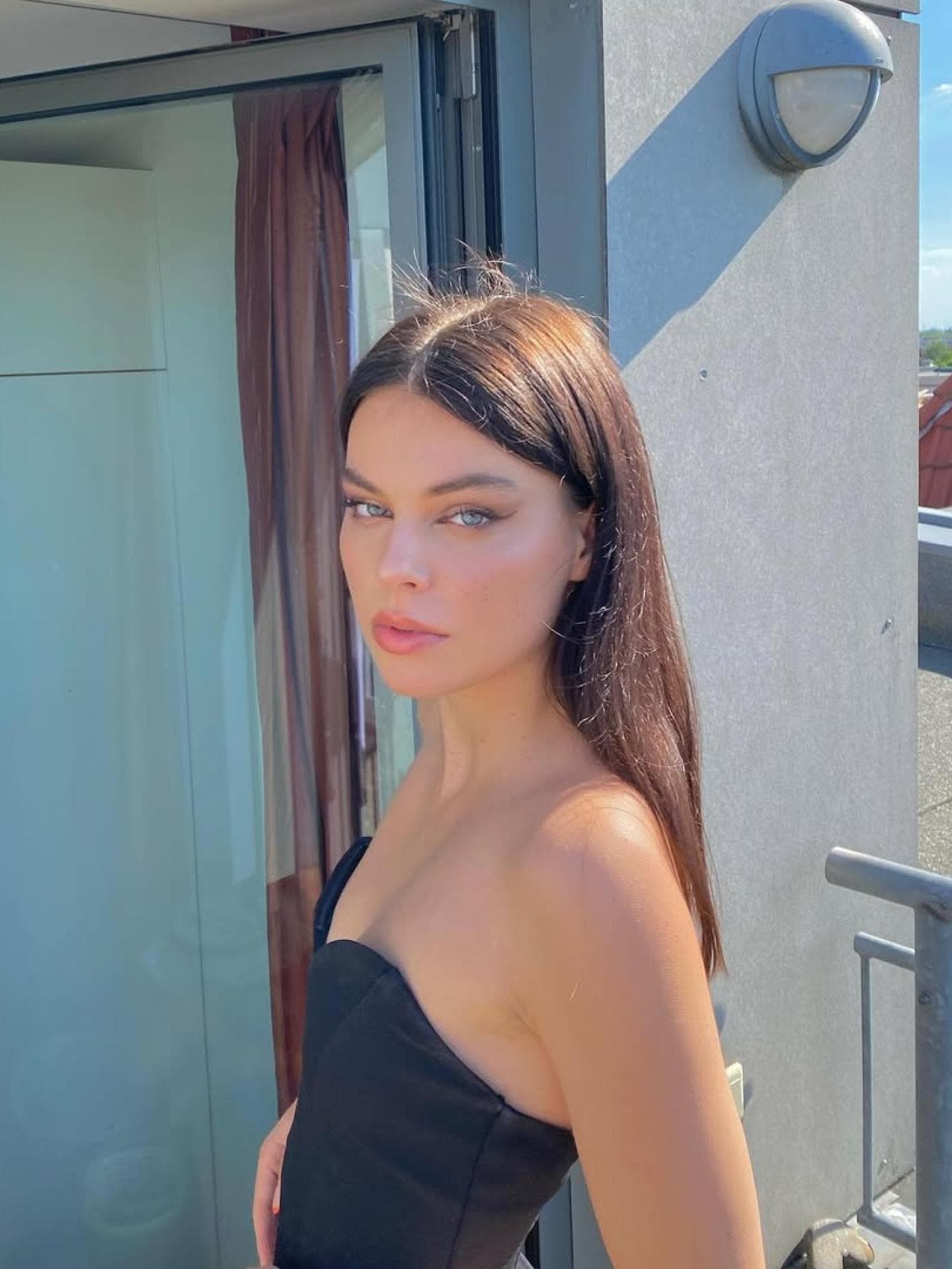 Selma hatte braune Haare, bevor sie bei GNTM teilnahm. Eine junge Frau mit braunen Haaren und einem trägerlosen Top auf einem Balkon in der Sonne.