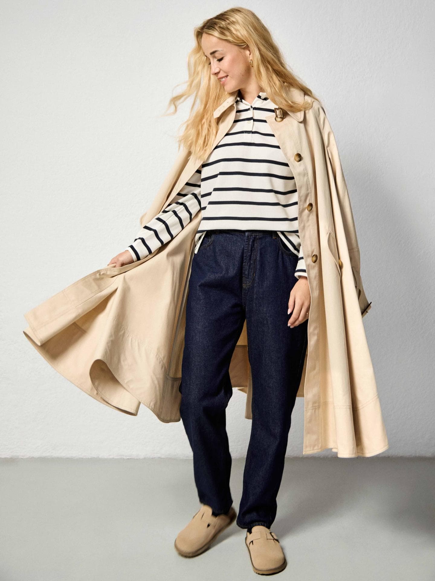 Head-to-toe-Look in TK Maxx: Ausgestellter Trenchcoat, Marinière im Polo-Stil, High-Waist-Jeans, Pantoletten, Ohrringe und Ringe von TK Maxx.