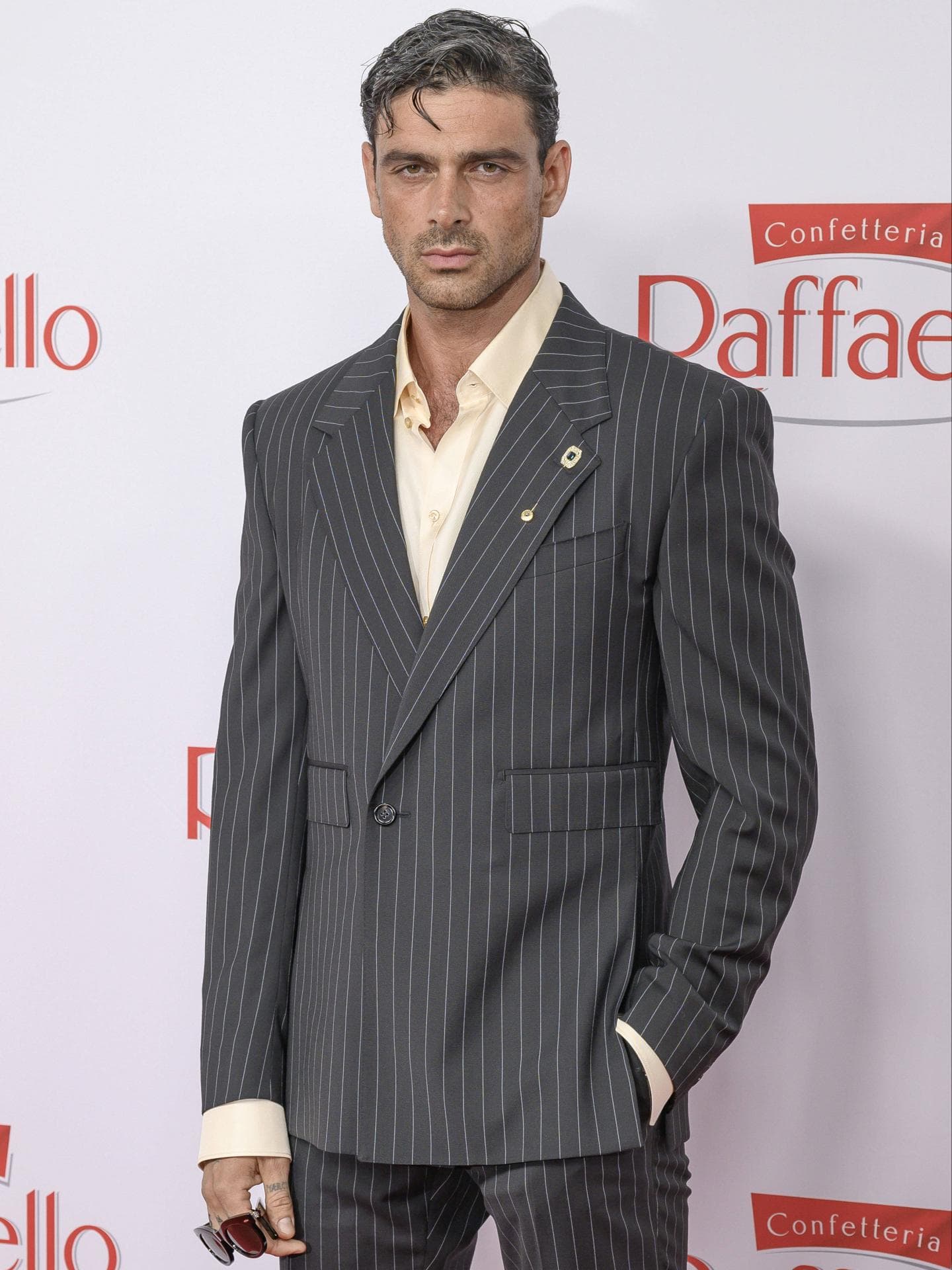 Zeitweise trug Michele Morrone keinen Bart. Michele Morrone mit einem grauen Anzug mit Nadelstreifen und einem hellgelben Hemd darunter.