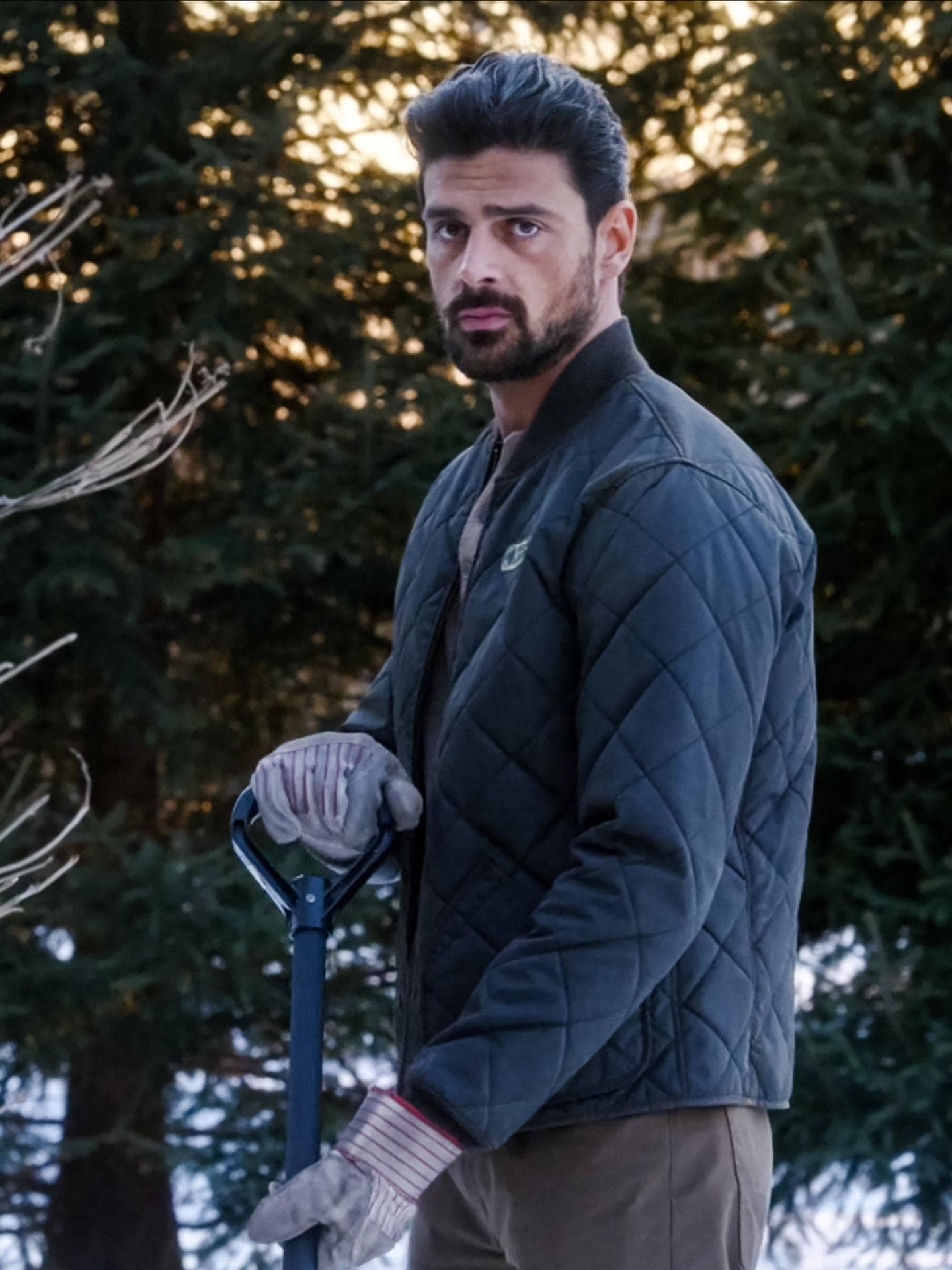 Michele Morrone in seiner Rolle als Gärtner in „The Housemaid“. Michele Morrone im Garten mit Handschuhen, einer Steppjacke und einem Sparten.