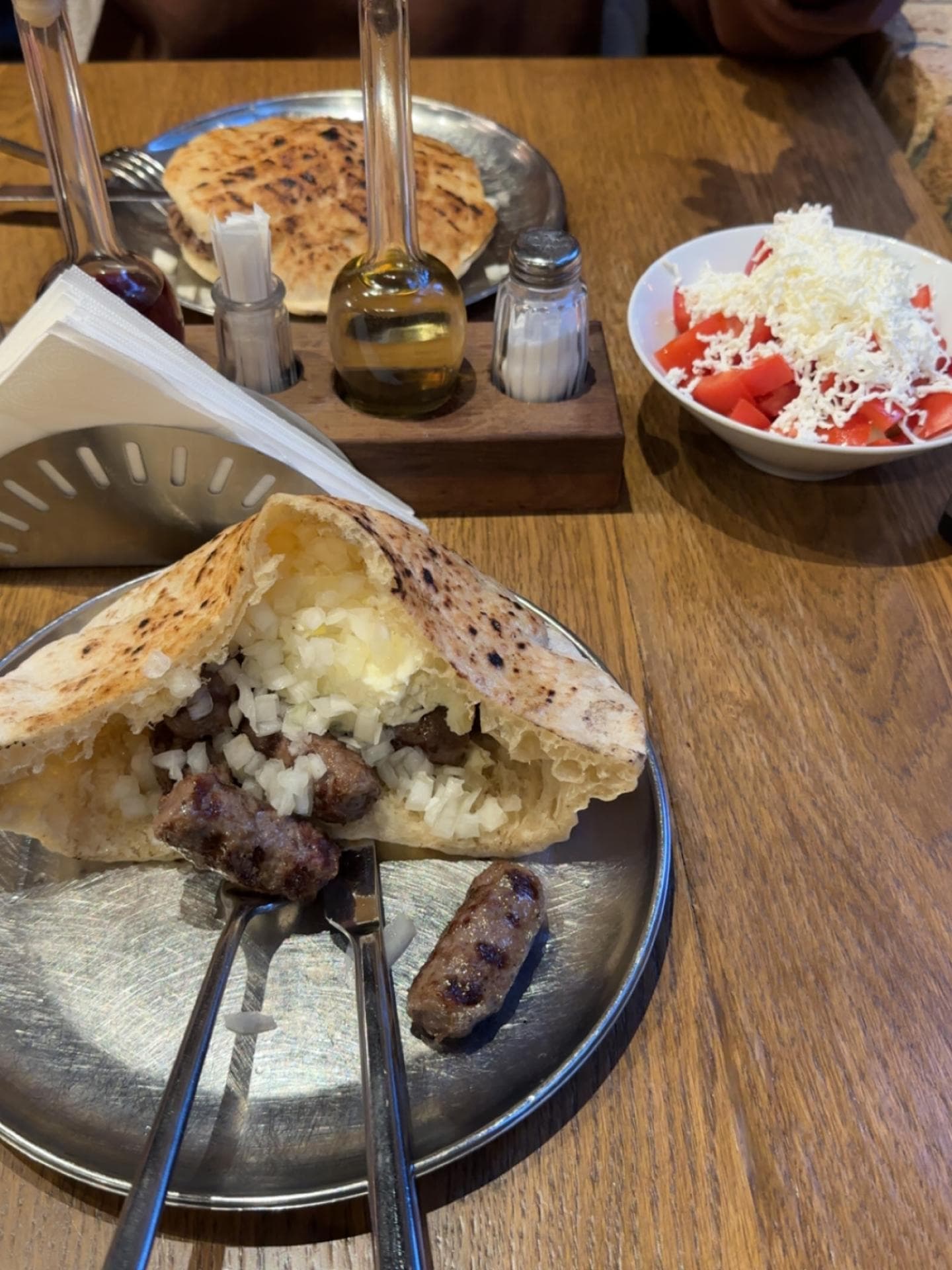 Cevapi im Brötchen mit Zwiebeln auf einem Silberteller auf einem Holztisch mit Salz, Pfeffer, Öl und einem Tomatensalat mit Käse.