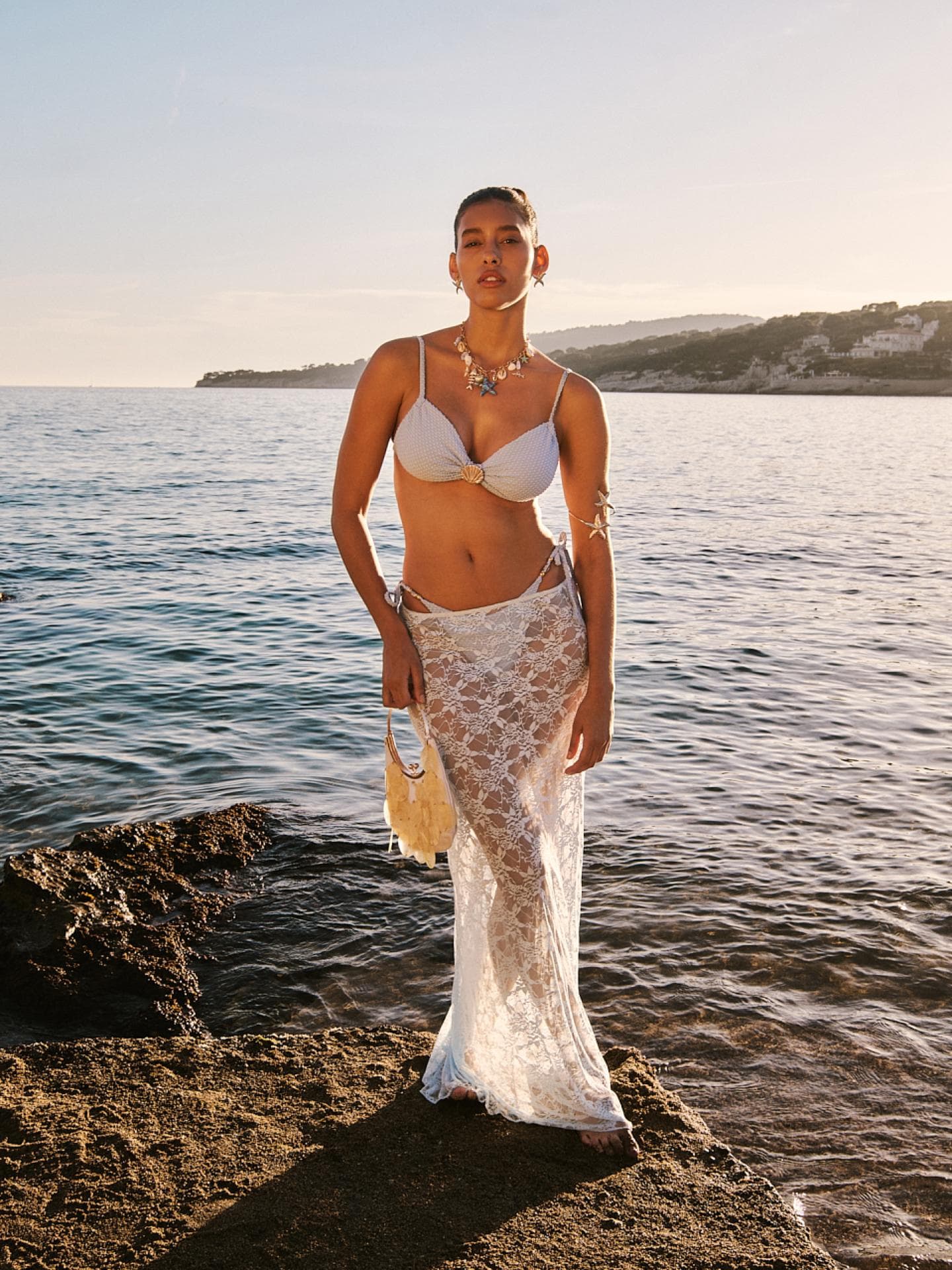 Eine junge Frau steht auf einem Felsen im Wasser und trägt einen weißen Bikini und einen Spitzenrock darüber.