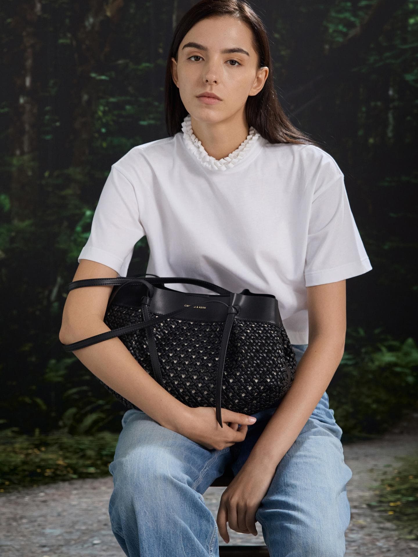 Die gewebte Tote Bag von Charles & Keith ist ein echter Hingucker zu jedem schlichten Outfit. Frau mit weißem Shirt und Jeans hat eine gewebte schwarze Tasche auf dem Schoß.