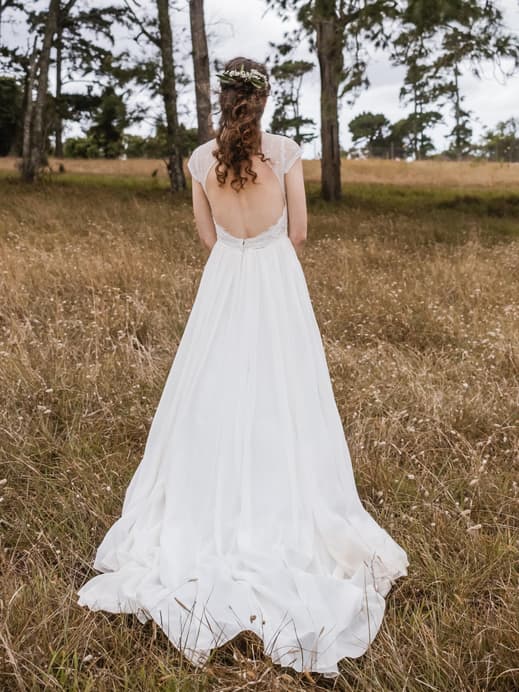 Frau mit Boho Hochzeitsfrisur steht auf einem Feld mit dem Rücken zur Kamera.