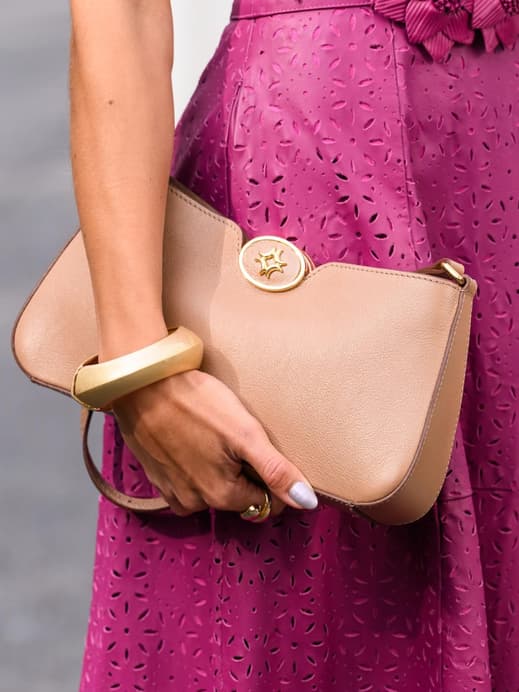 Eine Nahaufnahme einer Frau mit einem Kleid mit Lochmuster in Magenta und einer hellbraunen Clutch in der Hand mit goldenen Armreifen.