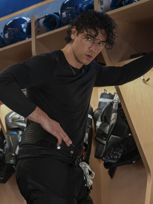 Ein junger Mann mit Locken steht in einem langärmeligen Shirt in der Umkleide einer Eishockey-Mannschaft.