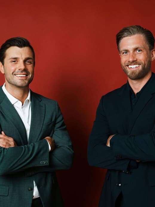 Tim Reitz und Sebastian Paul in Anzügen mit verschränkten Armen vor einer roten Wand.