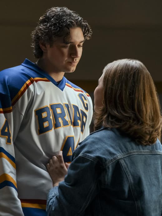 Ein junger Mann im Eishockey-Trikot steht einer jungen Frau in Jeansjacke gegenüber und guckt zu ihr herunter.