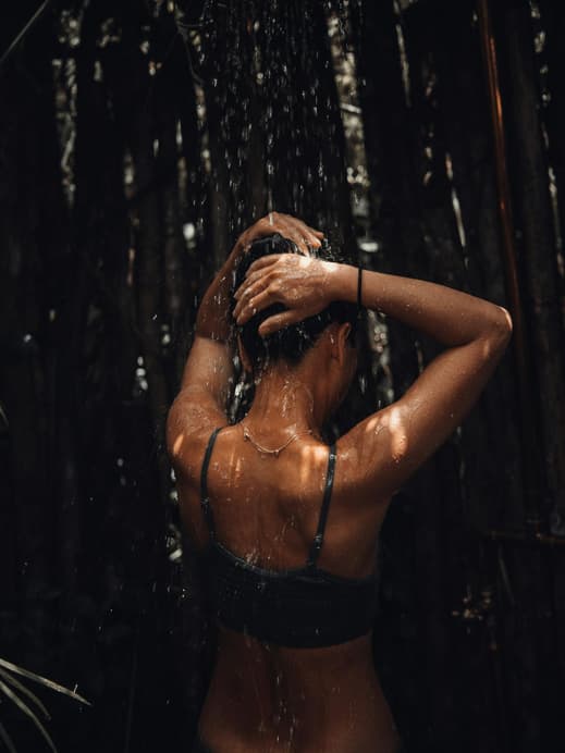 Eine Frau steht mit dem Rücken zur Kamera in einer Dusche und lässt sich mit den Armen über dem Kopf berieseln.