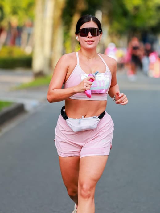 Eine junge Frau mit einer Sonnenbrille, Sportkleidung und einer kleinen Wasserflasche am Laufen auf der Straße.