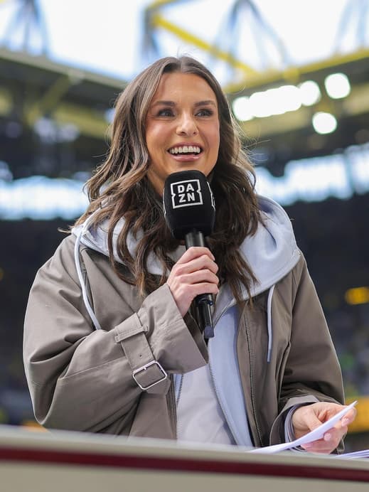 Eine junge Frau mit braunen Haaren steht in einem Fußballstadio und moderiert.