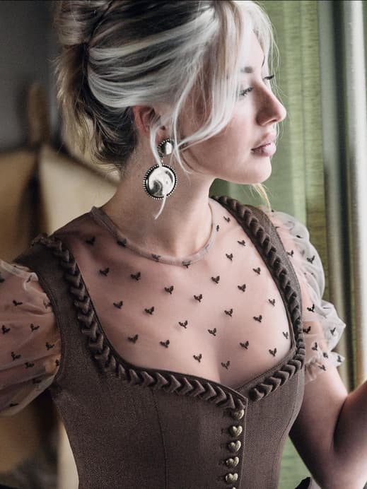 Frau mit hochgesteckten Haaren und Dirndl mit transparenter Bluse guckt durch ein Fester zur Seite.