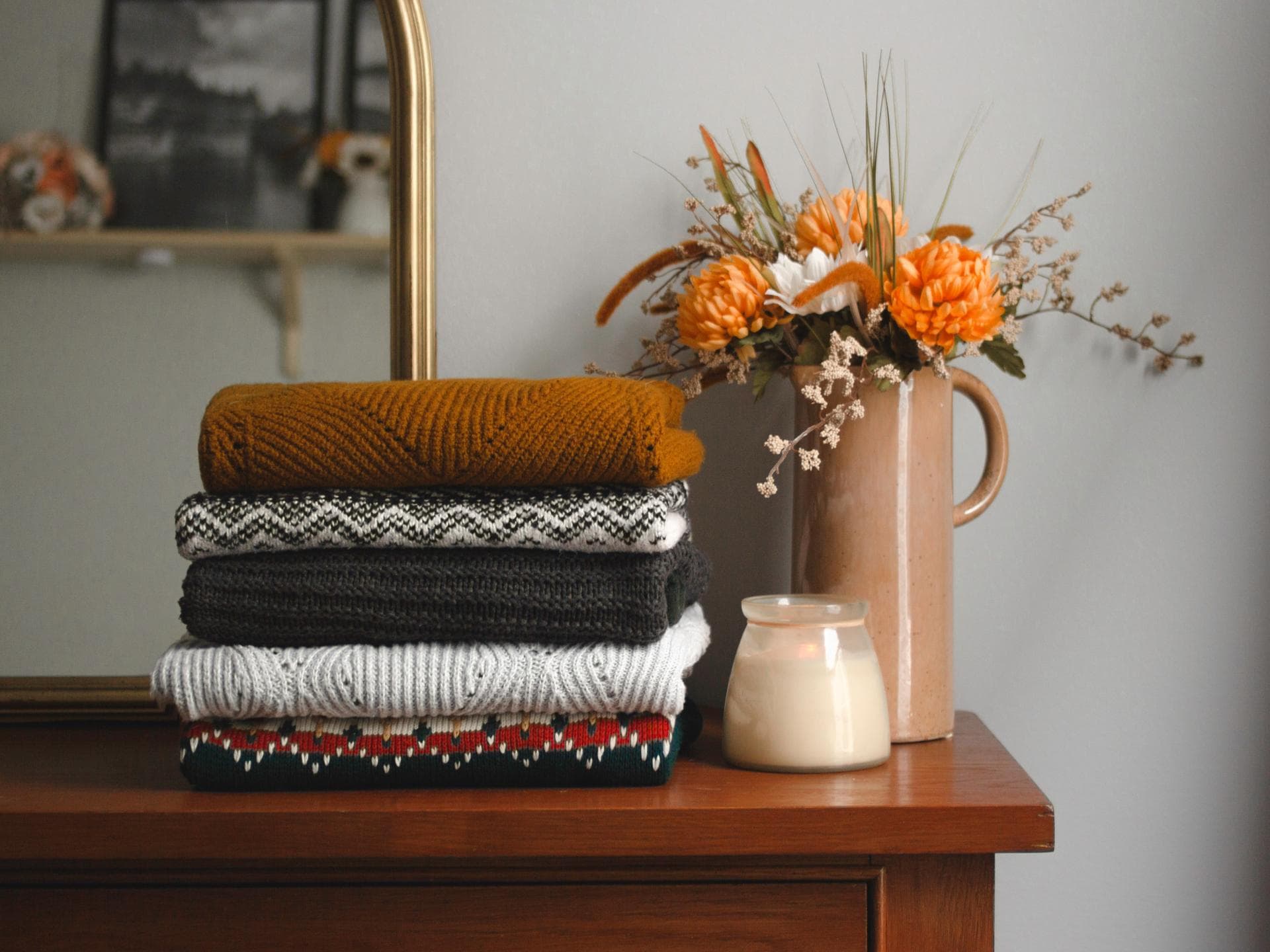 Gefaltete Wollpullover auf einer Holkommode neben einer Kerze und einer Blumenvase