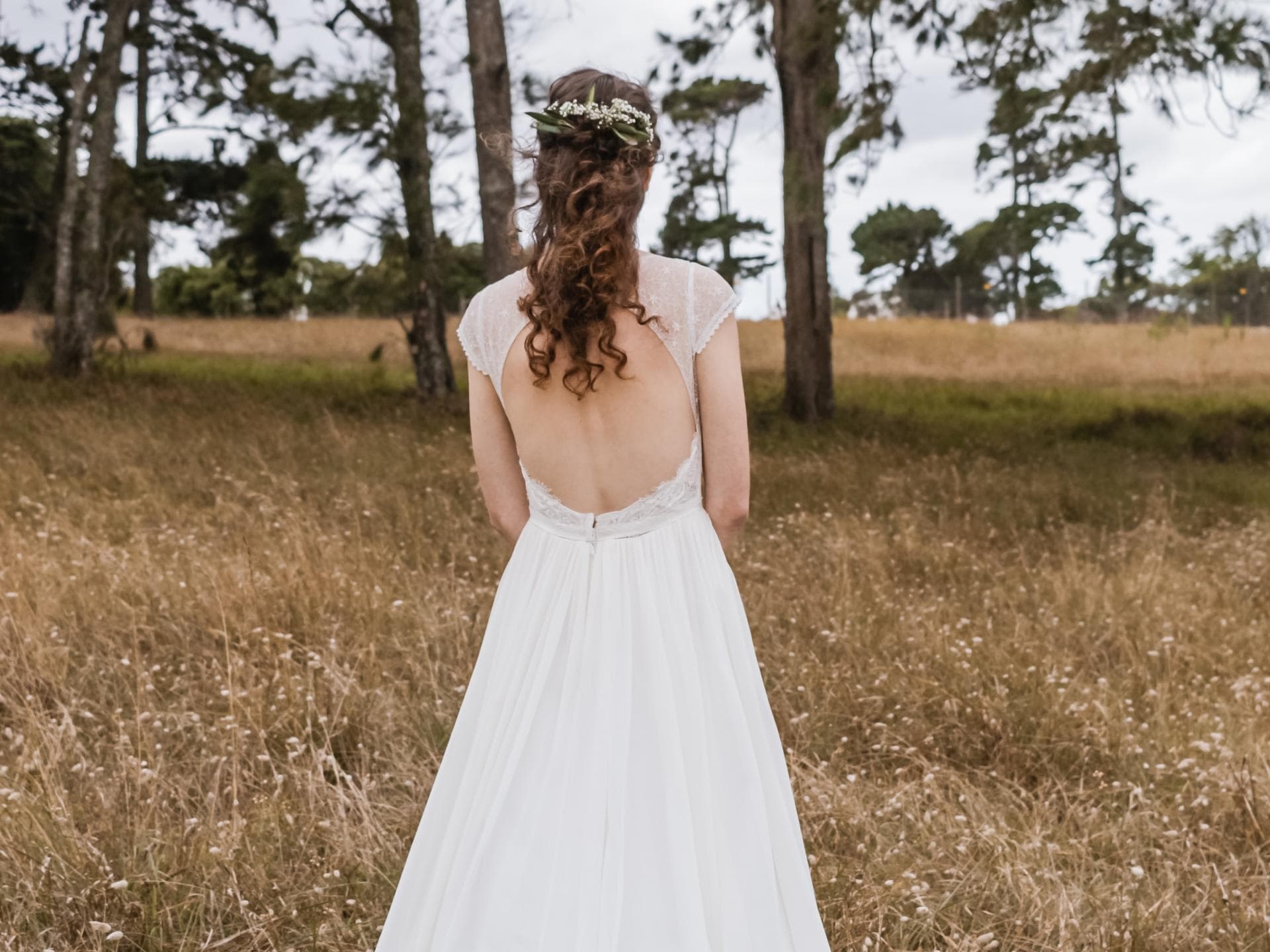 Frau mit Boho Hochzeitsfrisur steht auf einem Feld mit dem Rücken zur Kamera.