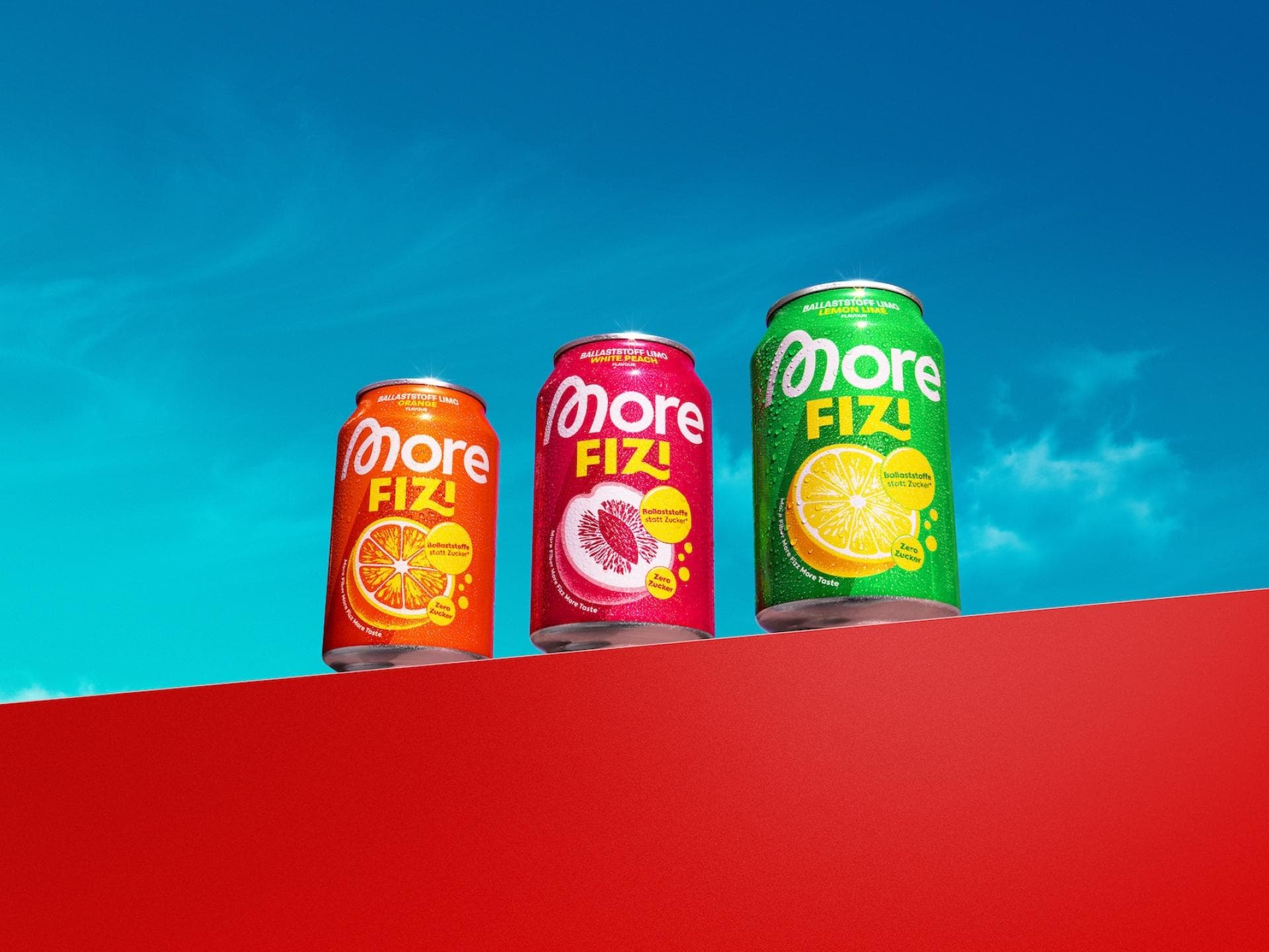 Drei bunte Getränkedosen mit der Aufschrift „More Fizi“ auf einer roten Mauer vor blauem Himmel.