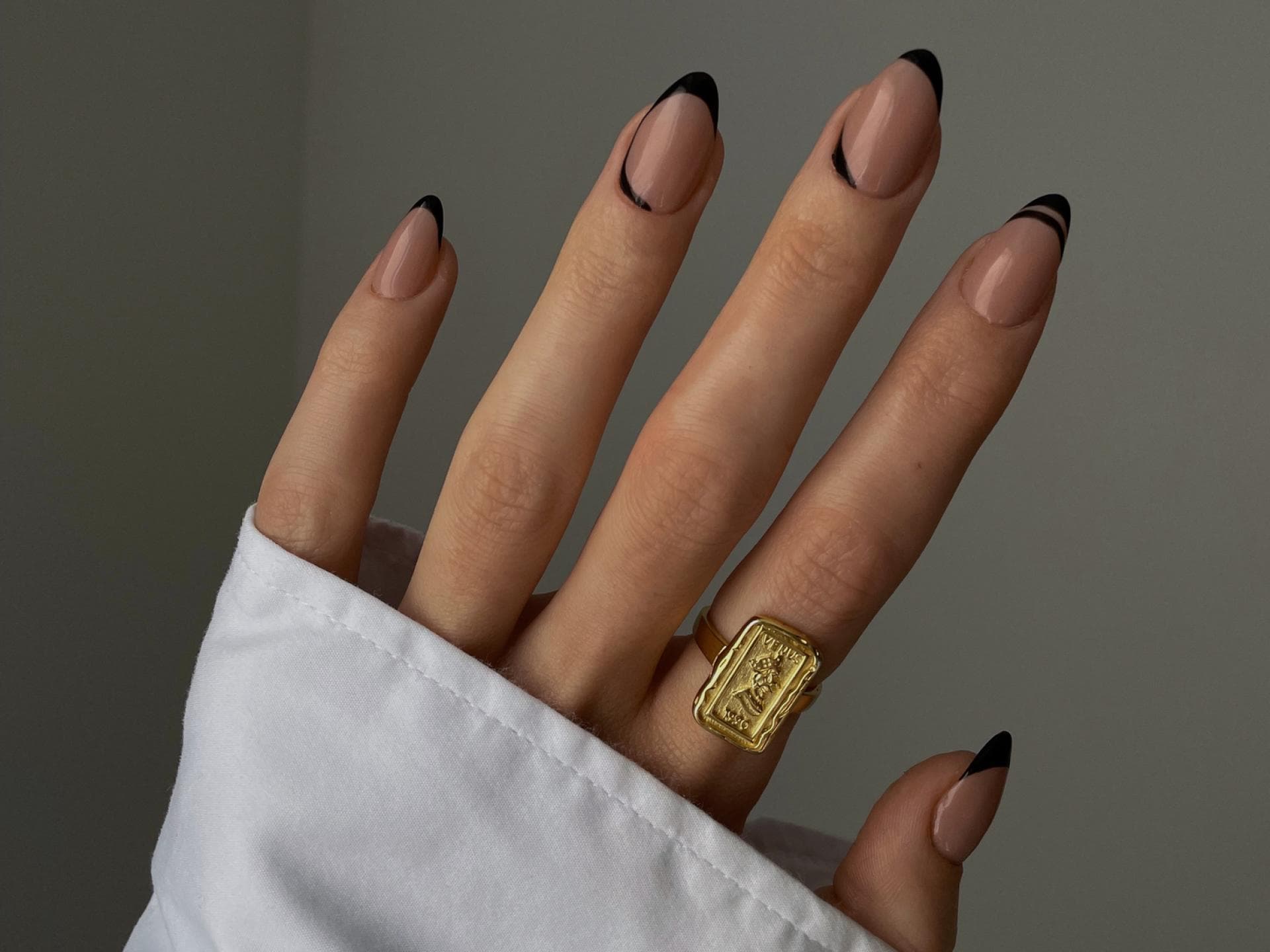 Curved French Nails liegen jetzt im Trend. Die Hand einer Frau mit schwarzen schwarzen French Nägeln und einem goldfarbenen Ring am Zeigefinger.